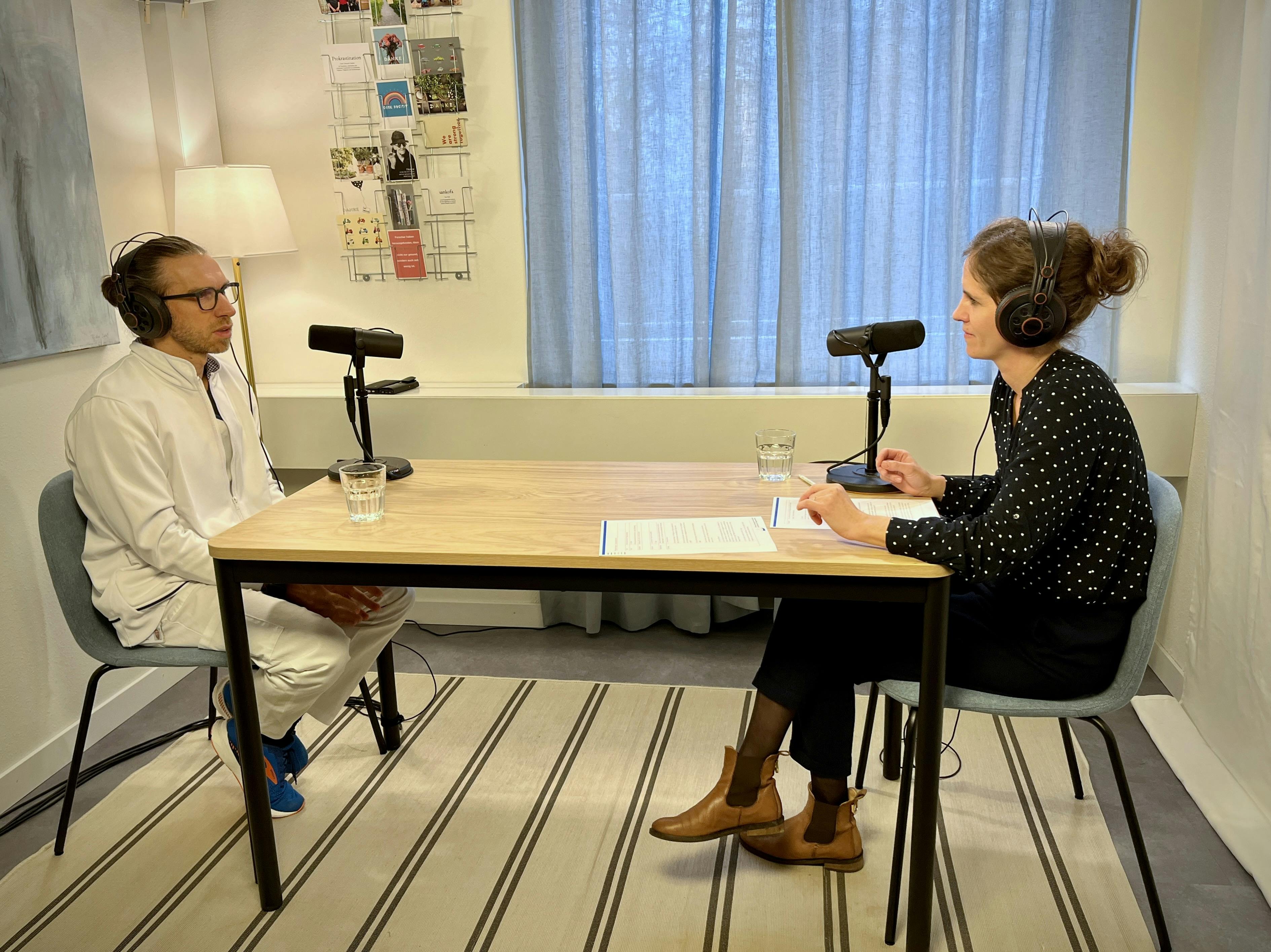 Zwei Personen bei einem Podcast-Interview in einem hellen Raum.